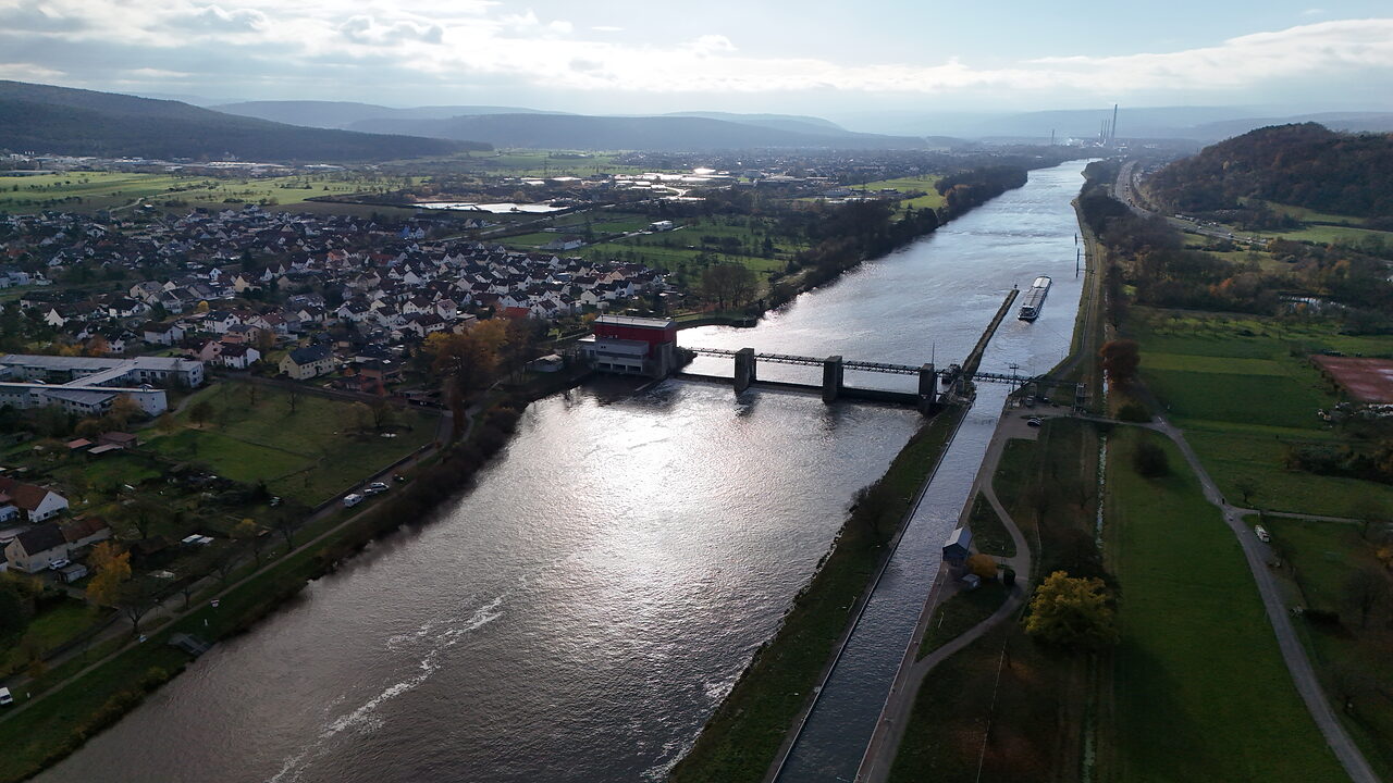 Luftaufnahme eines Flusses mit Staustufe, Ortschaft und Hügeln im Hintergrund